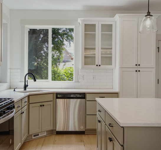 View into a new kitchen with island, upper and lower cabinets and kitchen sink positioned in a corner under two windows.